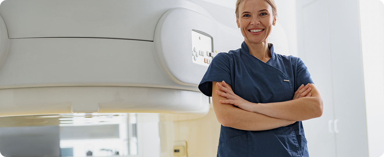 Smiling female healthcare professional in blue scrubs standing with arms crossed beside medical imaging equipment.