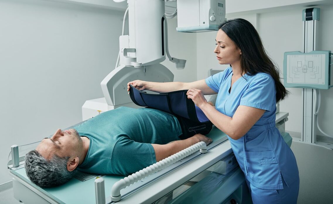 Medical professional in blue scrubs preparing a male patient for an X-ray procedure while he lies on a radiology exam table.